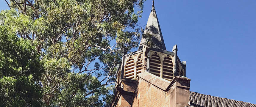 Photograph of the spire and upper section of St John's Church at Gordon Cemetery, with ornate architectural details. The building is partially obscured by tall trees, and the sky is clear and blue.