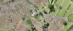 Aerial view of Northern Metropolitan Cemeteries showing a grid of burial plots, pathways, and green spaces. Roads divide different sections of the cemetery, and a small building is visible in the center of the image, surrounded by trees and greenery.