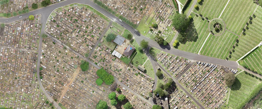 Aerial view of Northern Metropolitan Cemeteries showing a grid of burial plots, pathways, and green spaces. Roads divide different sections of the cemetery, and a small building is visible in the center of the image, surrounded by trees and greenery.