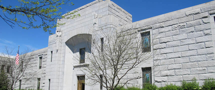 Photograph of a stone building at Ferncliff Cemetery, featuring large, light gray stone blocks and arched windows. A leafless tree stands in front of the building, with an American flag visible on the left side, and the sky is clear and blue.