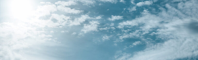 Photograph of a bright blue sky with scattered white clouds, with the sun partially visible in the top left corner, creating a soft glow