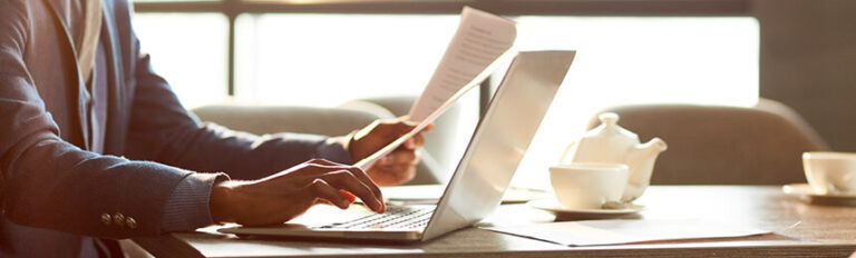 Photograph of a person working on a laptop while holding a document, sitting at a table with a teapot, teacup, and other papers in a softly lit room.