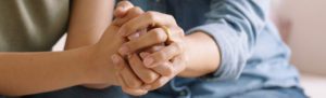 Close-up photograph of two people holding hands in a gesture of support and comfort, with their fingers interlocked.