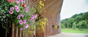 Photograph of a building at Green Acres Cemeteries and Memorial Parks, featuring a wooden exterior with hanging baskets of pink and yellow flowers. The building is set against a backdrop of green trees and an open landscape.