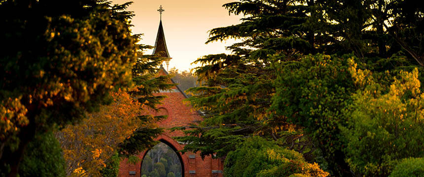 Photograph of a red brick chapel with a steeple at Carr Villa Cemetery, partially obscured by tall trees and lush greenery. The soft golden light of sunset adds warmth to the scene.